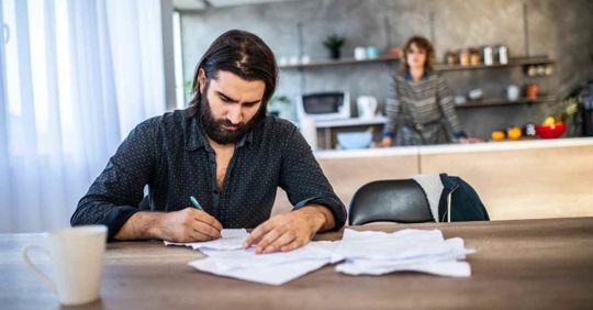 A man sitting at a kitchen table filling out paperwork. His wife stands in the background looking at him